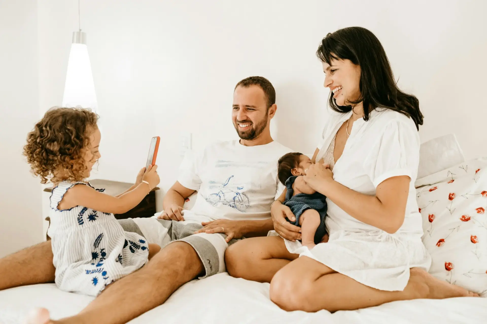 Eltern mit Kleinkind und Baby sitzen entspannt zusammen auf dem Bett: Familienalltag mit zwei Kindern